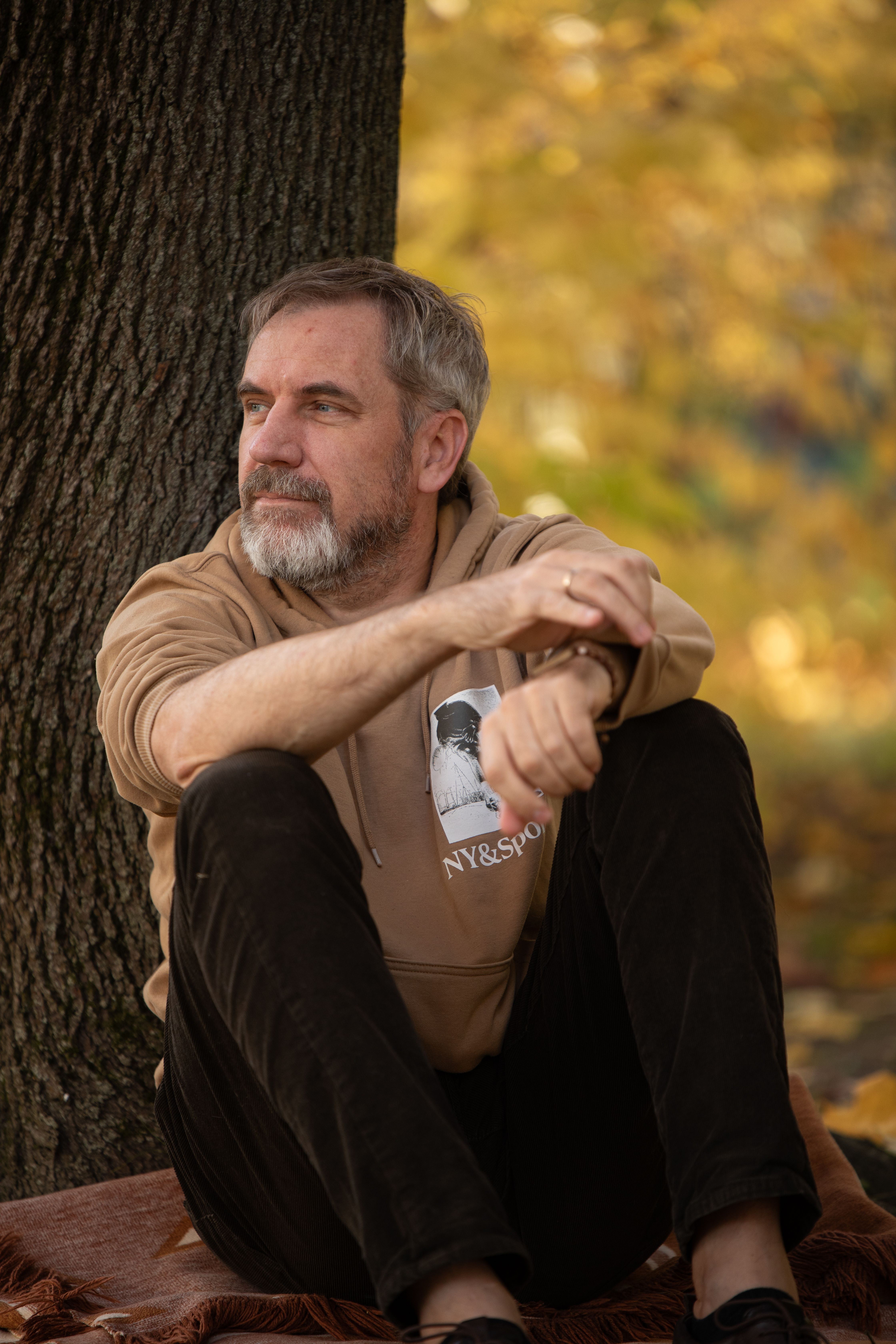 Pensive adult male in a hoodie sits against a tree trunk amidst fall foliage.