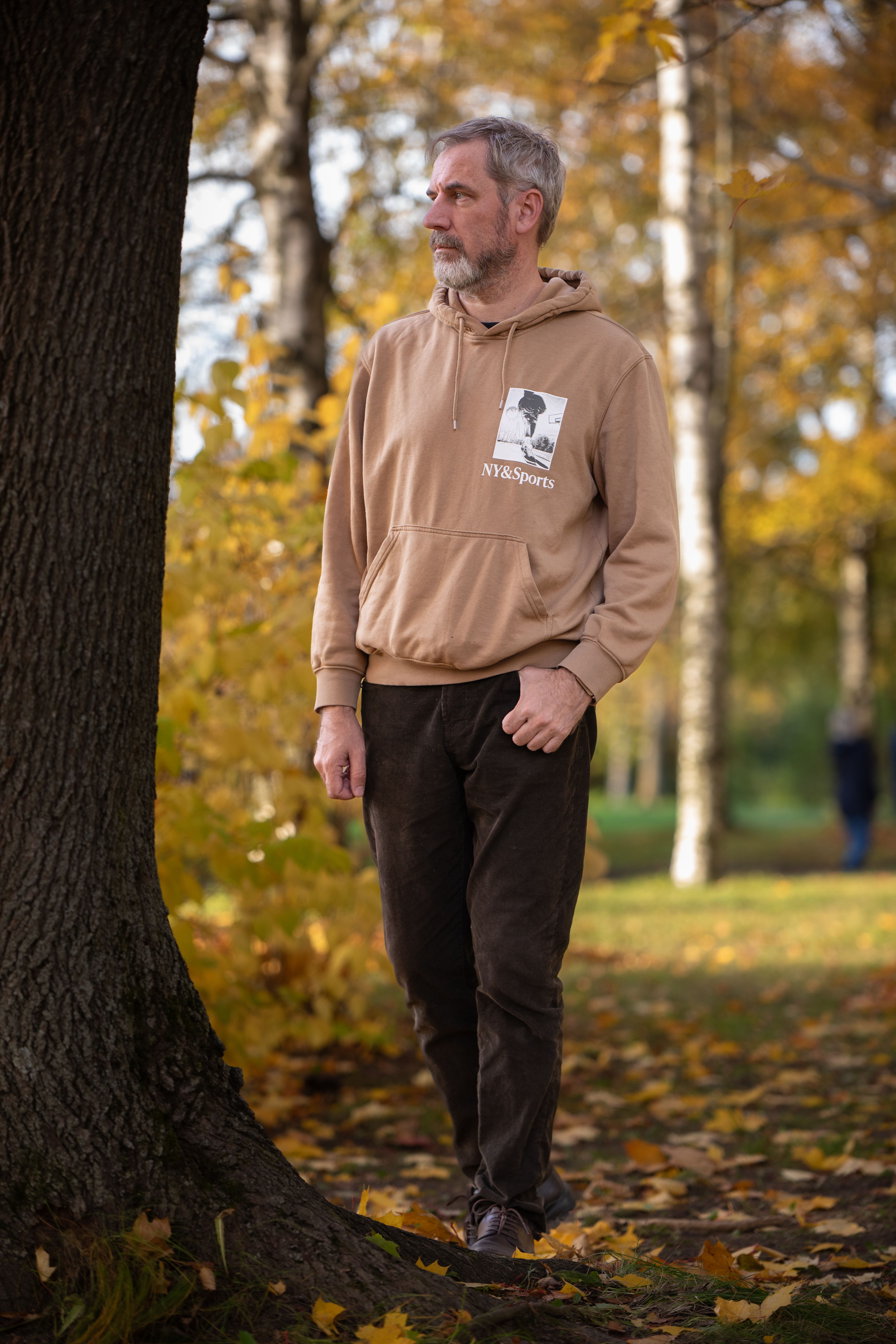Man walking by a tree in the park during fall, wearing a casual hoodie. Autumn leaves on the ground.