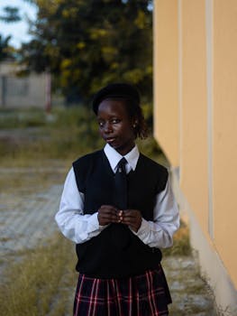 A young woman stands confidently in a school uniform next to a yellow building, displaying a thoughtful expression.
