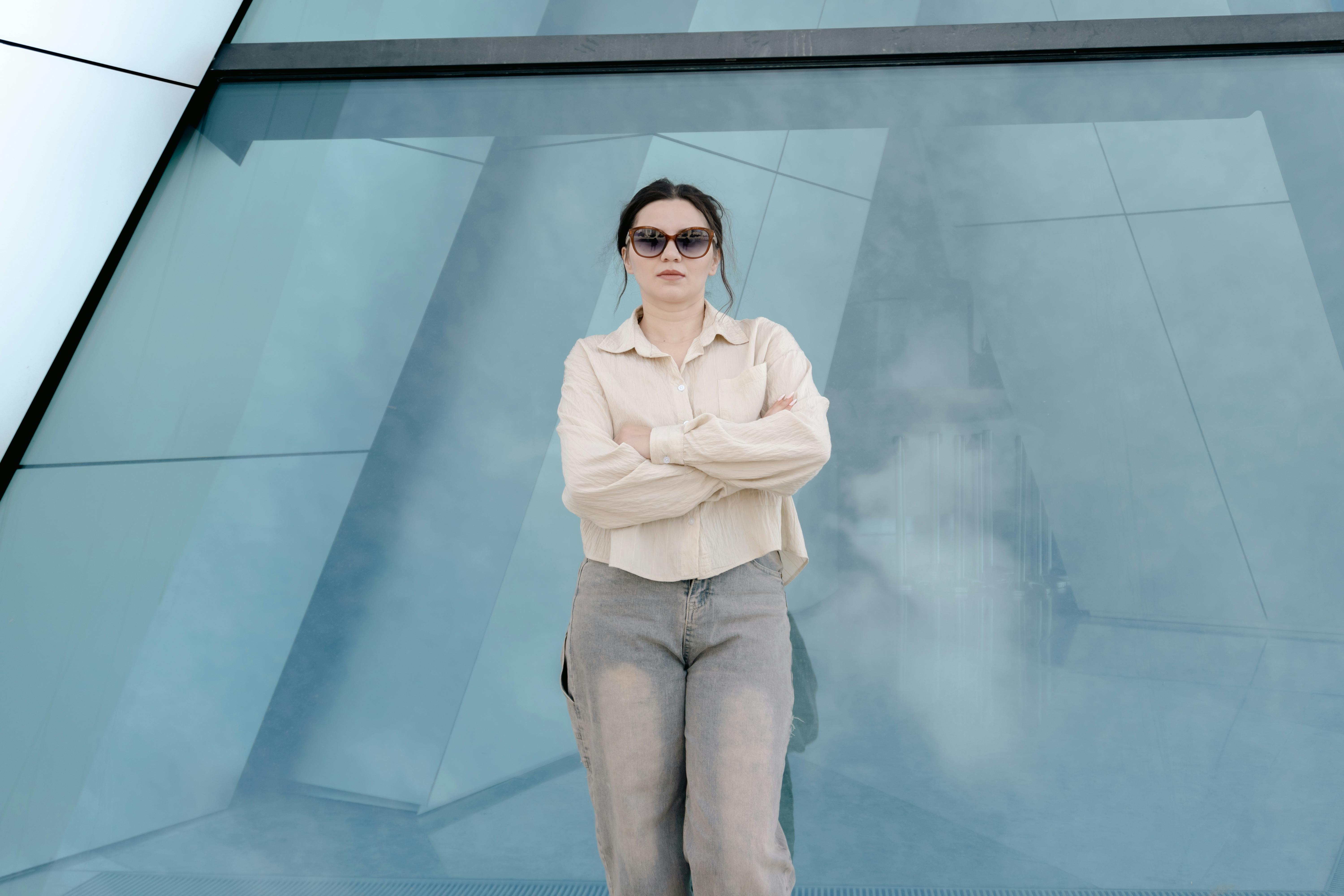 grátis Uma mulher estilosa, de óculos escuros, está confiante, com os braços cruzados, em frente a uma arquitetura moderna e elegante. Foto profissional