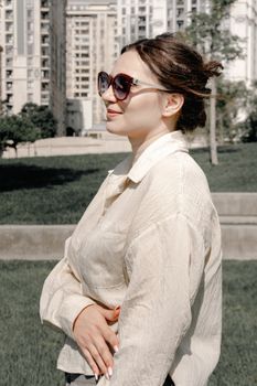 Stylish woman with sunglasses stands confidently outdoors against urban backdrop.