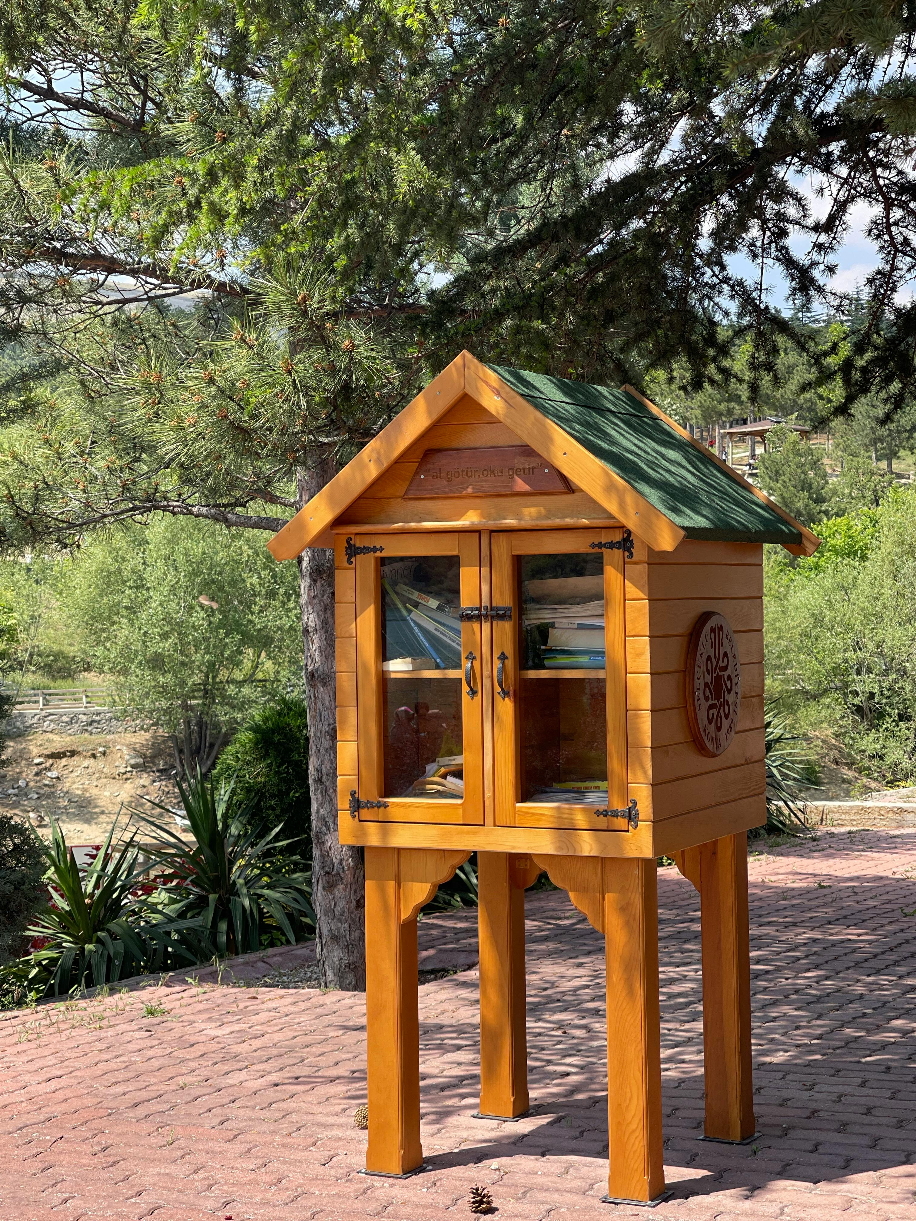 A cozy wooden little free library outdoors surrounded by greenery in daylight.