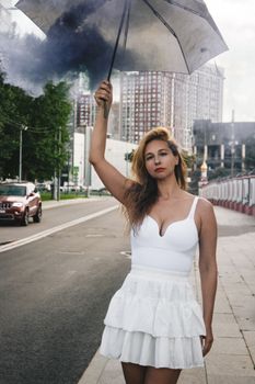 Woman in white dress holding an umbrella with smoke on a city street.