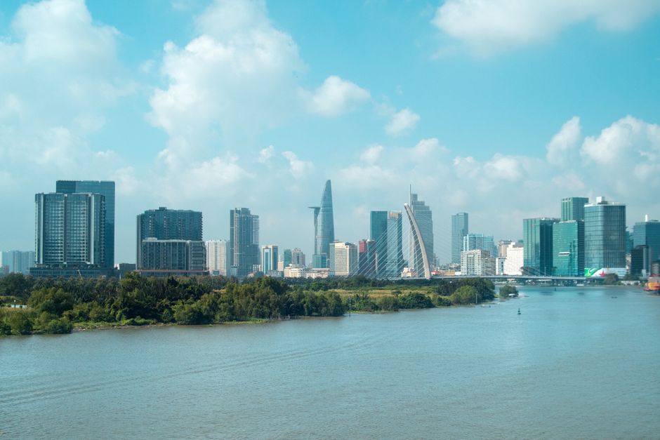 Vibrant Ho Chi Minh City skyline view with skyscrapers and river foreground under a bright blue sky.