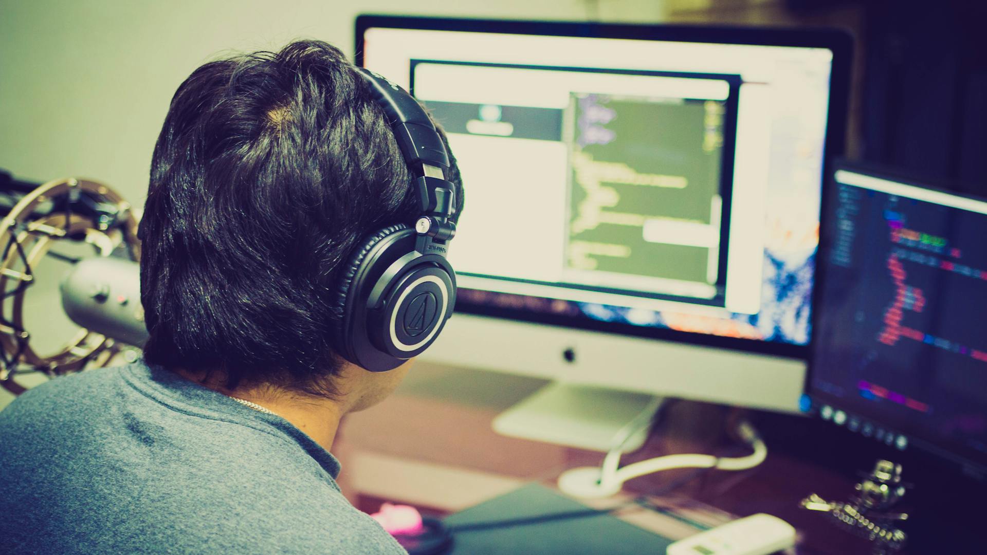 A programmer with headphones focuses on coding at a computer setup with dual monitors.