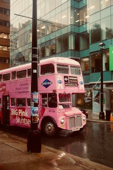 Vintage pink double-decker bus in a rainy urban setting, showcasing city life.