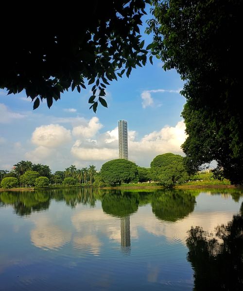 Free A modern tower reflects in a tranquil lake surrounded by lush trees on a sunny day. Stock Photo