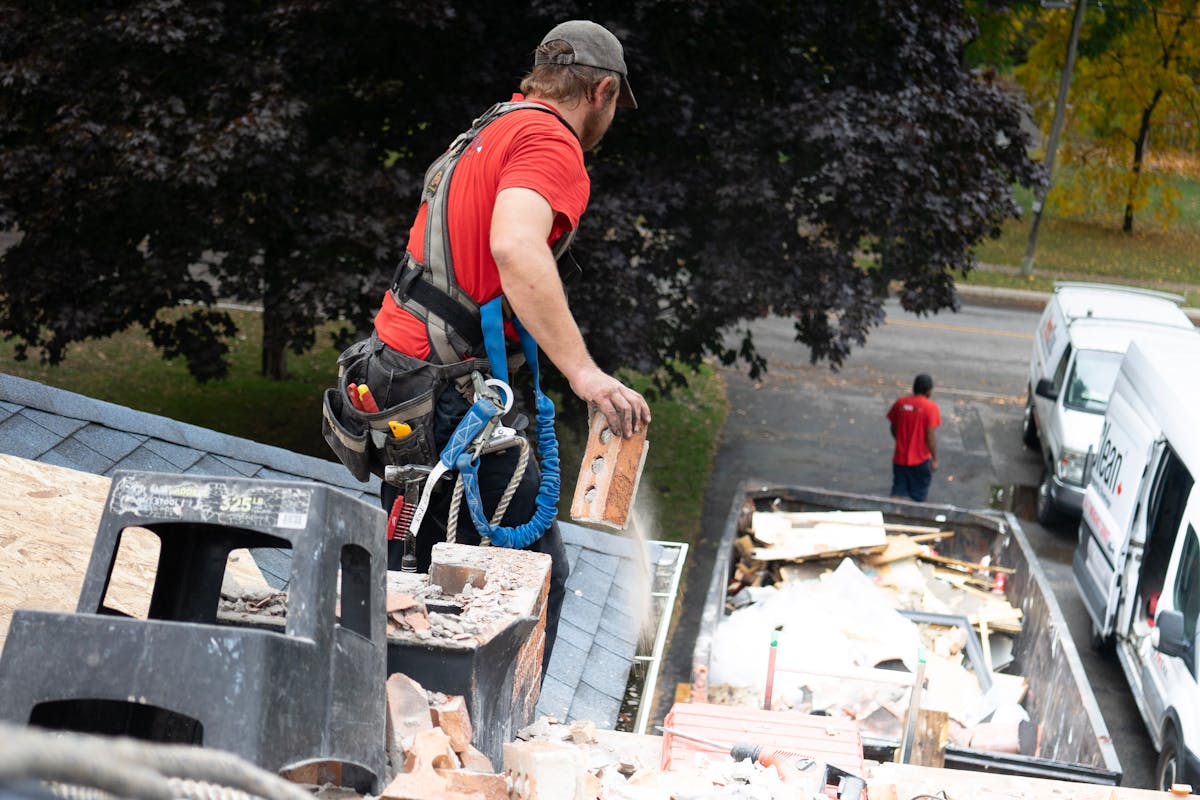 Professional roofer in safety harness removing debris from roof into dumpster demonstrating proper property protection and organized jobsite management with specialized equipment