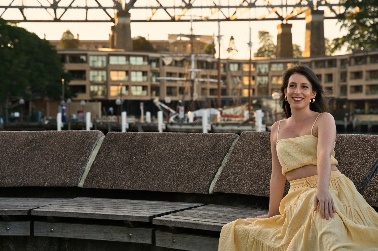 Smiling woman in a yellow sundress sits on a waterfront bench in warm evening light