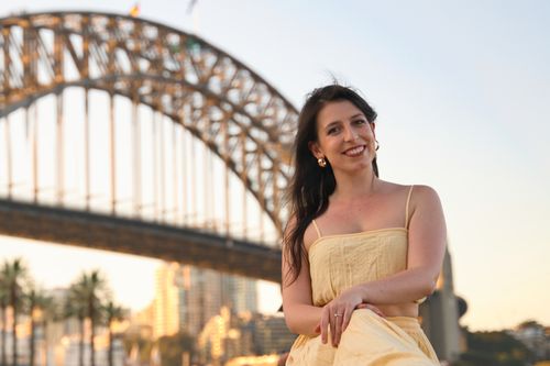 Free A cheerful woman poses in front of the iconic Sydney Harbour Bridge during sunset, creating a memorable moment. Stock Photo