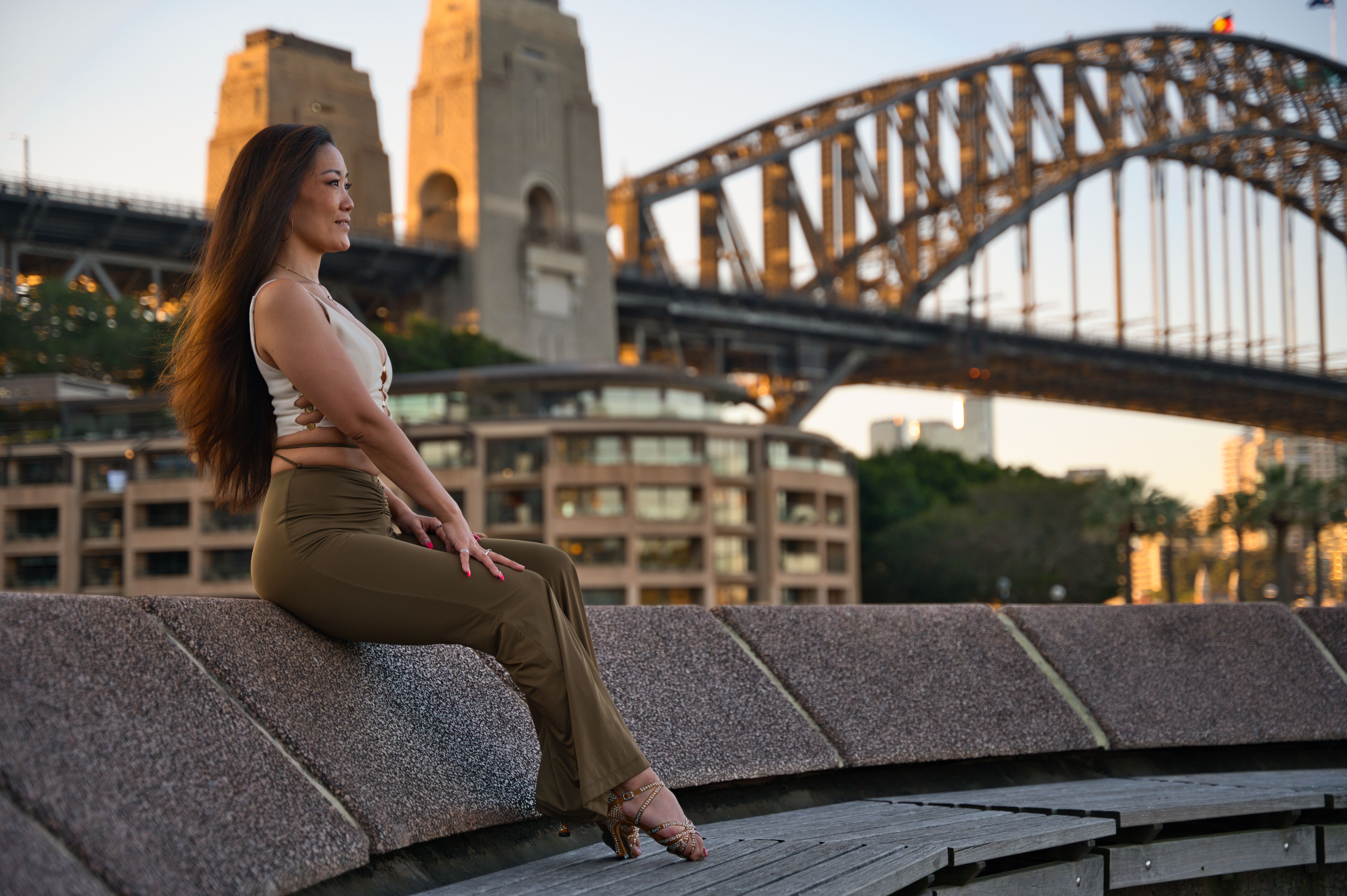 A woman sits elegantly near the iconic Sydney Harbour Bridge as the sun sets, capturing a serene moment.