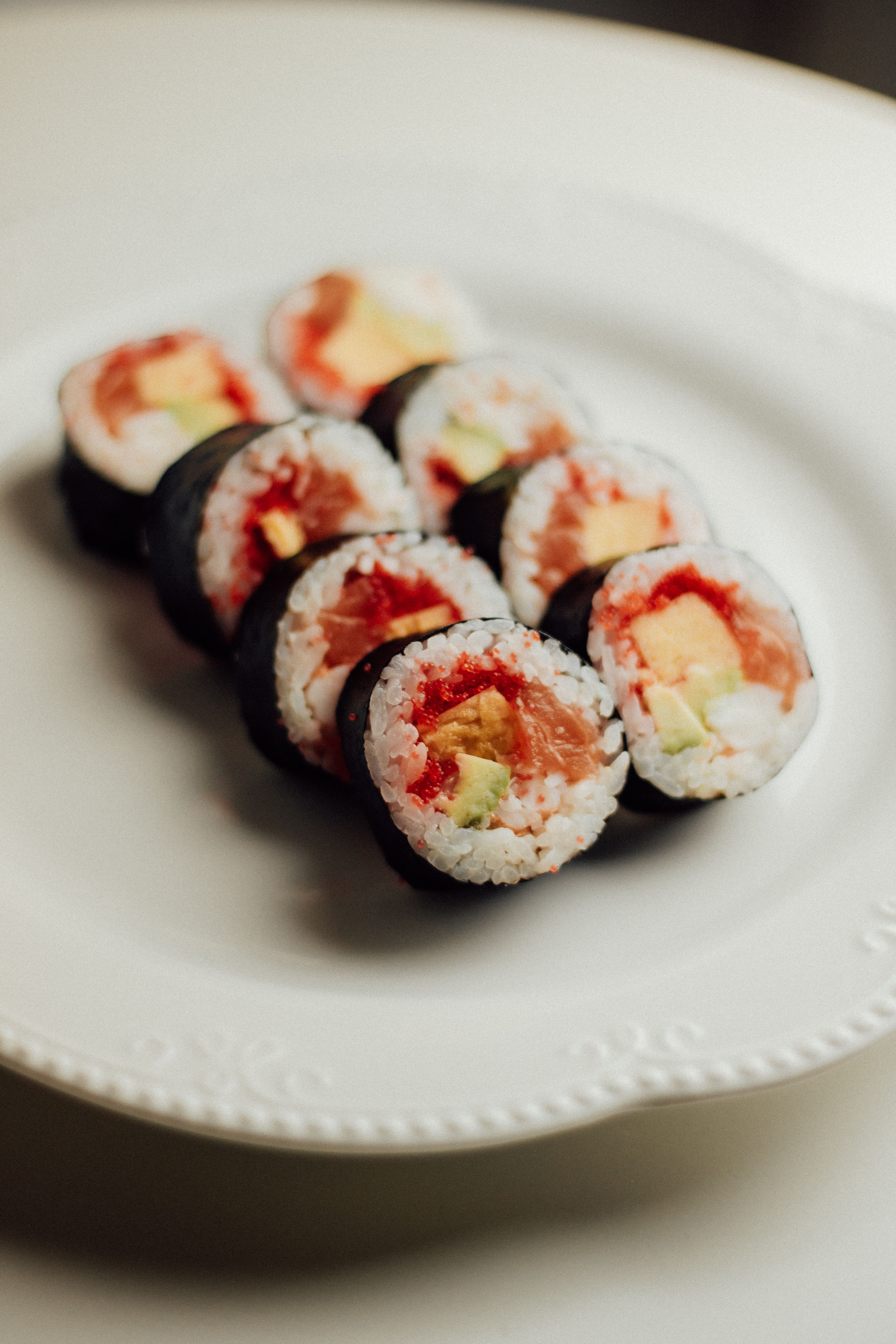 Sliced Alaska roll sushi pieces topped with a thin layer of salmon and avocado, showing the cucumber and rice layers inside, arranged on a plate with pickled ginger and wasabi.