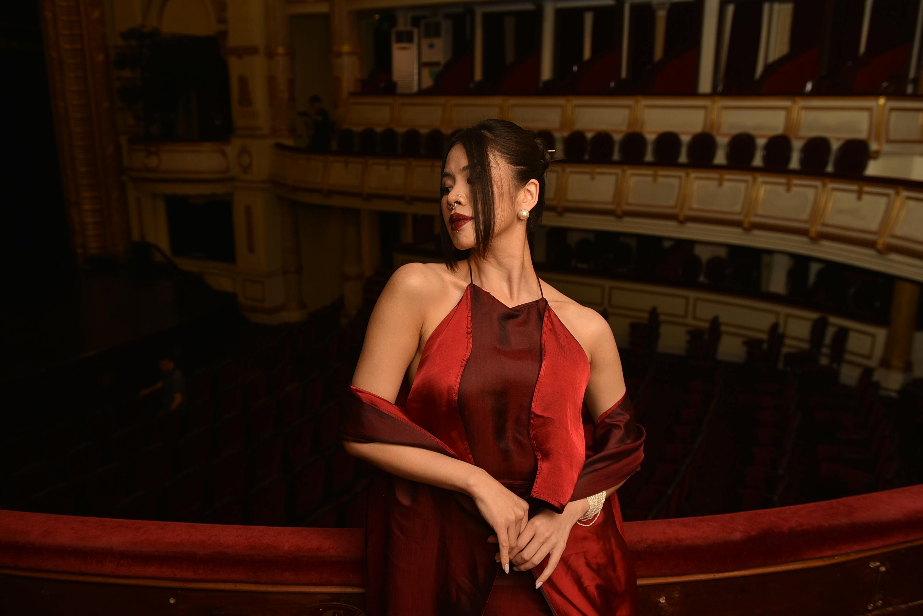 grátis Mulher estilosa em um elegante vestido vermelho posando na sacada de um teatro, exalando sofisticação. Foto profissional