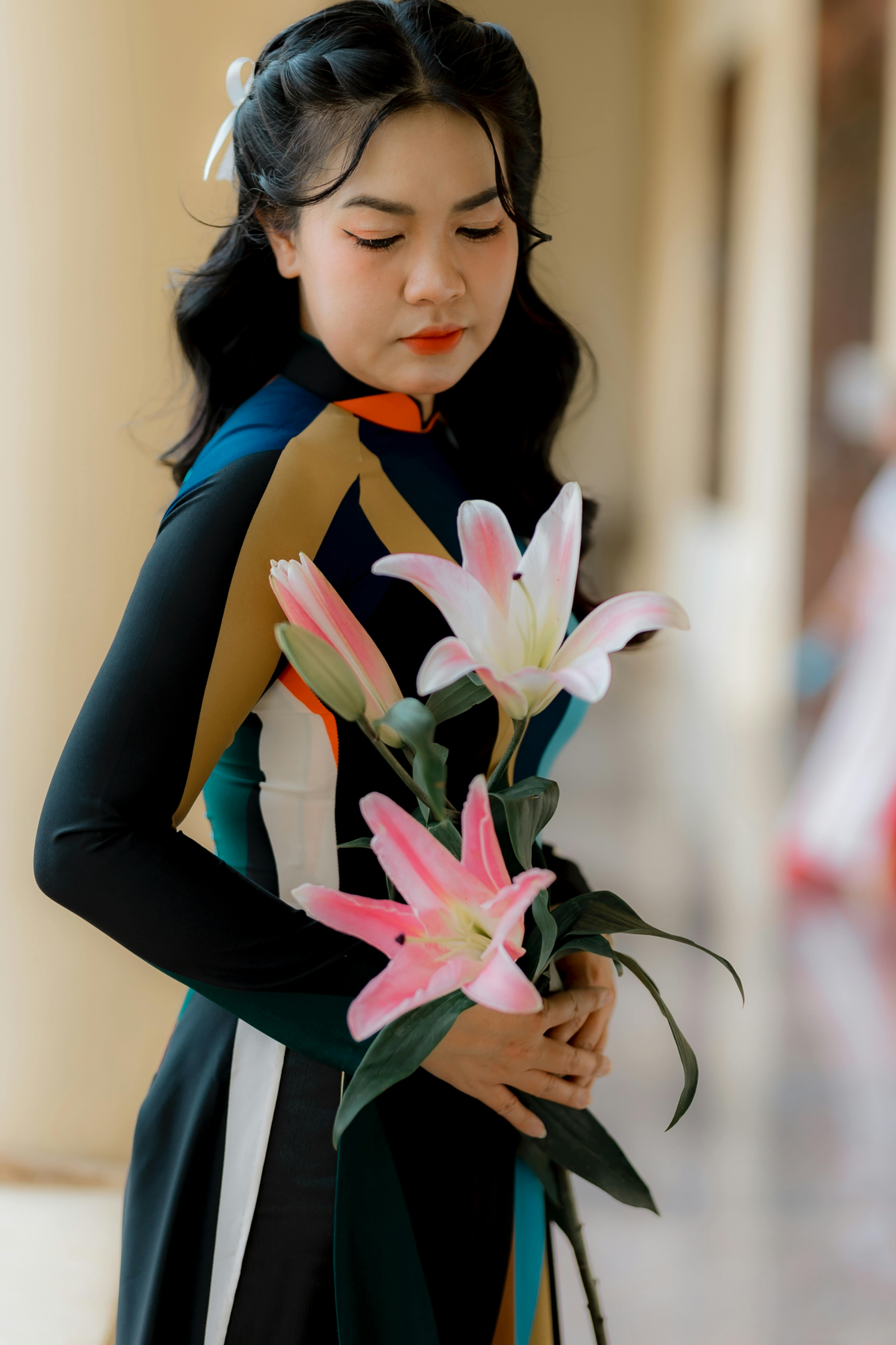 A young woman in traditional dress holds a bouquet of pink lilies inside a building.