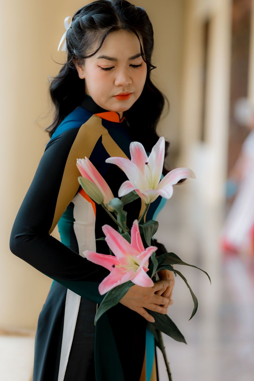 A young woman in traditional dress holds a bouquet of pink lilies inside a building.