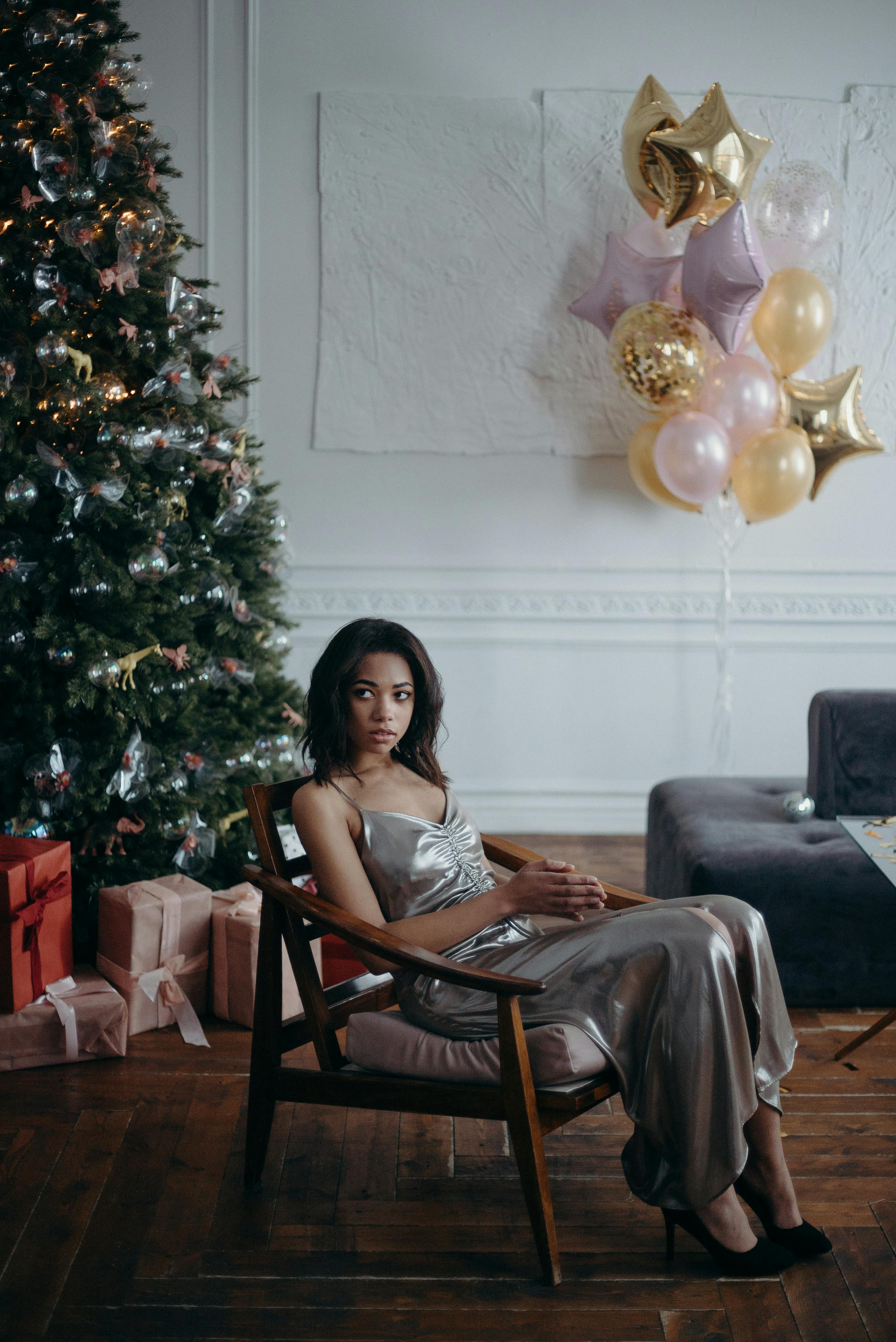 一位身穿银色连衣裙的女子坐在圣诞树旁，周围是点缀着礼物和气球的节日装饰