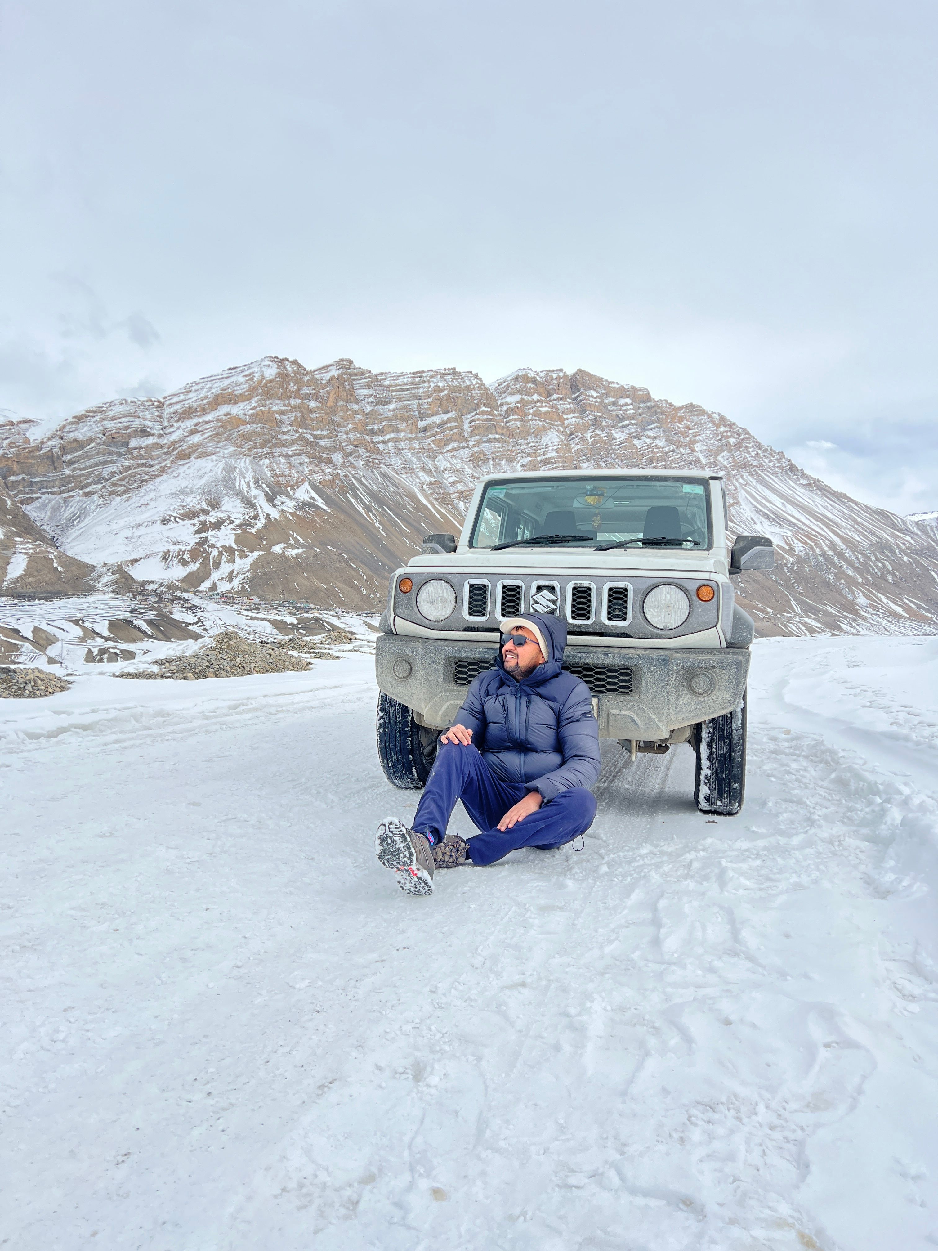 Man sitting by 4x4 vehicle on snowy road in Spiti Valley, showcasing winter adventure scenery.