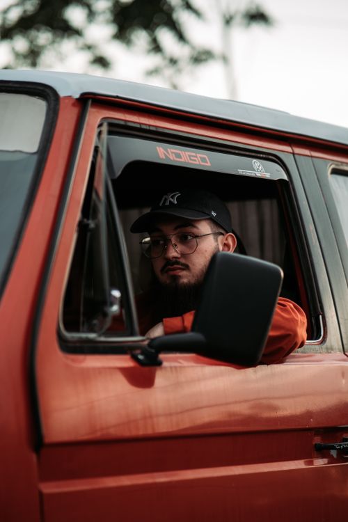 Free Portrait of a young man in a red van, wearing glasses and a cap, enjoying a drive outdoors. Stock Photo