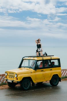 A woman in sunglasses poses on a yellow vintage jeep by the seaside under a cloudy sky.