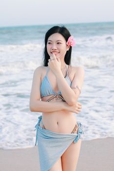 Smiling woman in blue bikini with pink flower on beach, enjoying sunny day.
