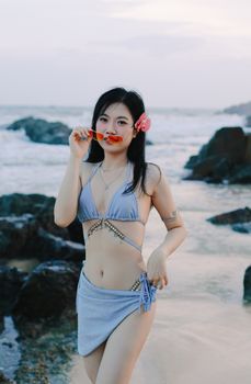 Young woman in blue bikini with sunglasses and flower on beach at sunset.