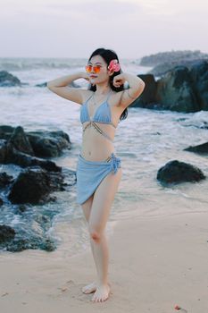 Woman in a blue bikini poses on a rocky beach, exuding a relaxed and summery vibe.