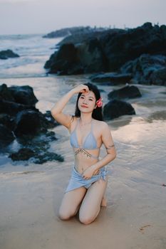 Woman in blue bikini kneeling on sandy beach with rocky shore and ocean waves at sunset, enjoying summer vibe.