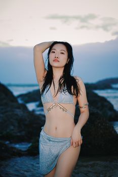 A woman poses confidently on the rocky shoreline at sunset, showcasing beachwear fashion.