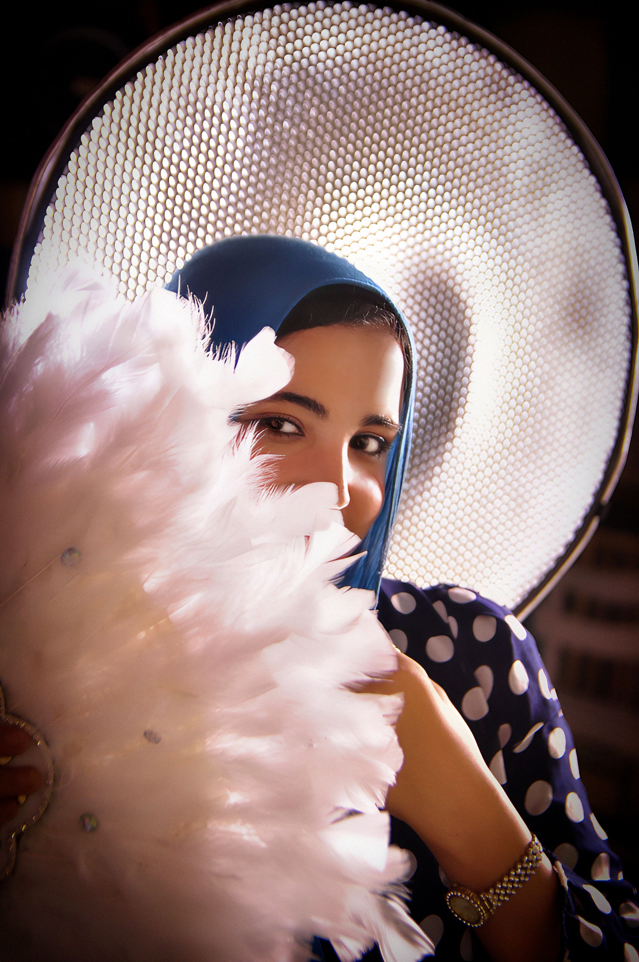 Elegant portrait of a woman in a blue hijab holding a feathered fan, with artistic lighting effect.