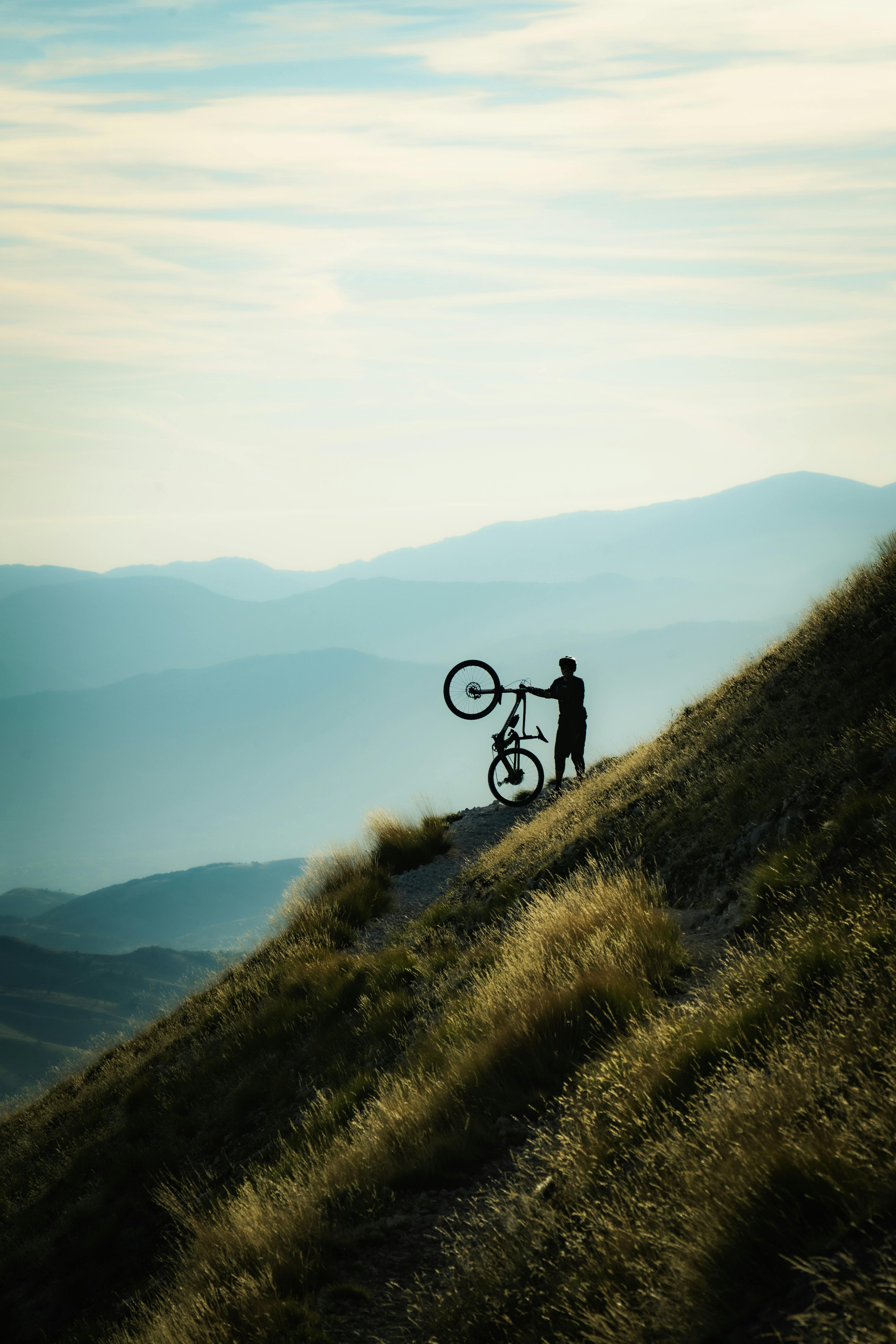 A lone cyclist stands on a mountain path, silhouetted against a twilight sky.