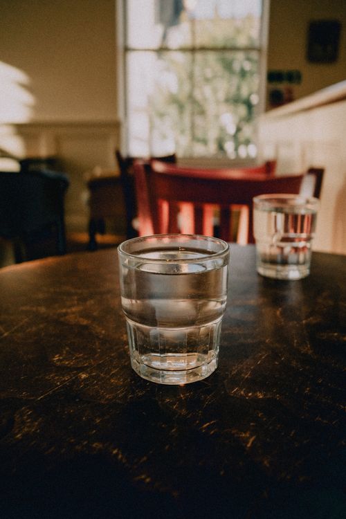 Free Warm sunlight illuminating a glass of water on a wooden table in a cozy café setting. Stock Photo