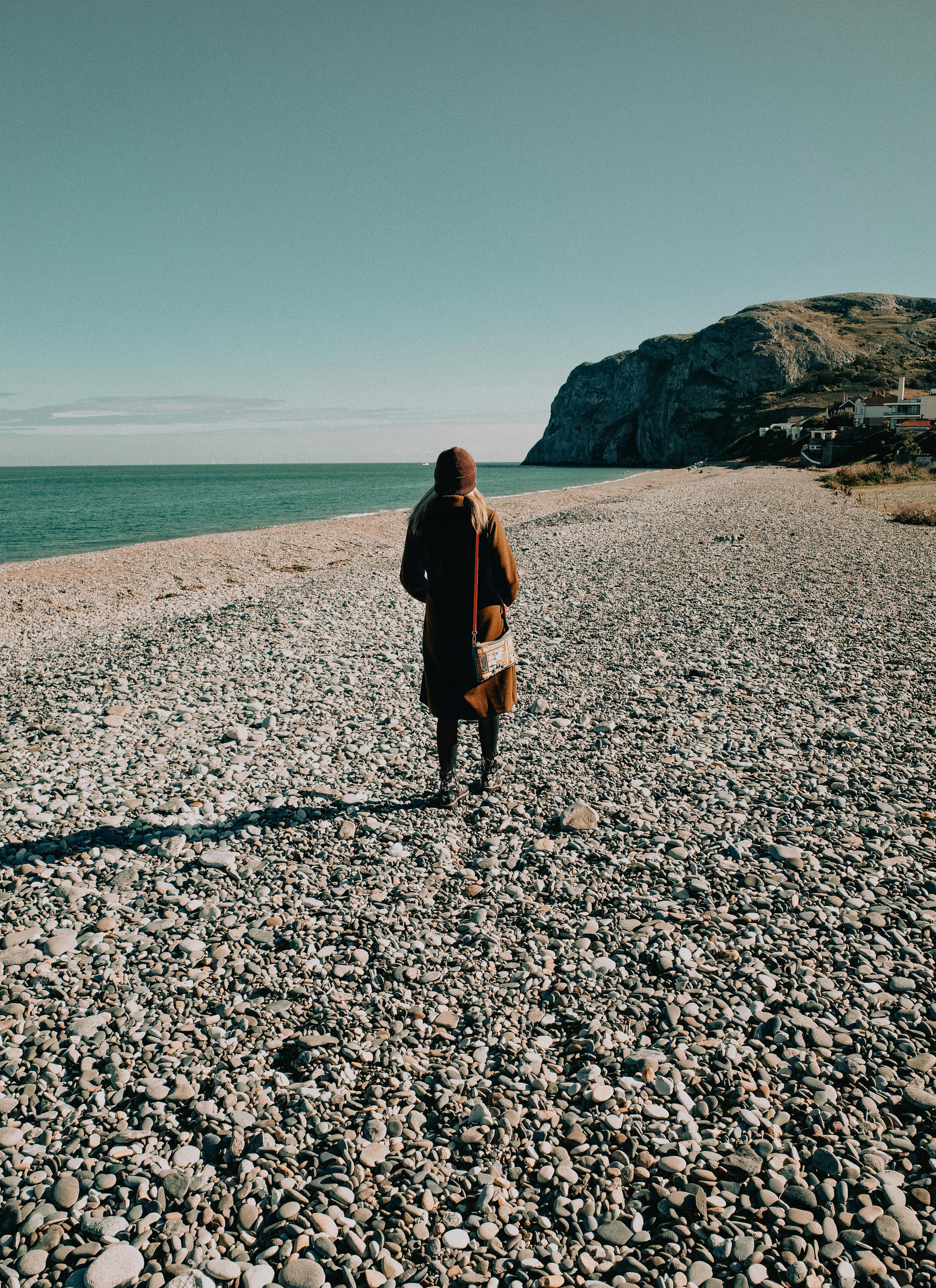 A solitary figure walks along a pebble beach in Llandudno, Wales, with a distant cliff view.