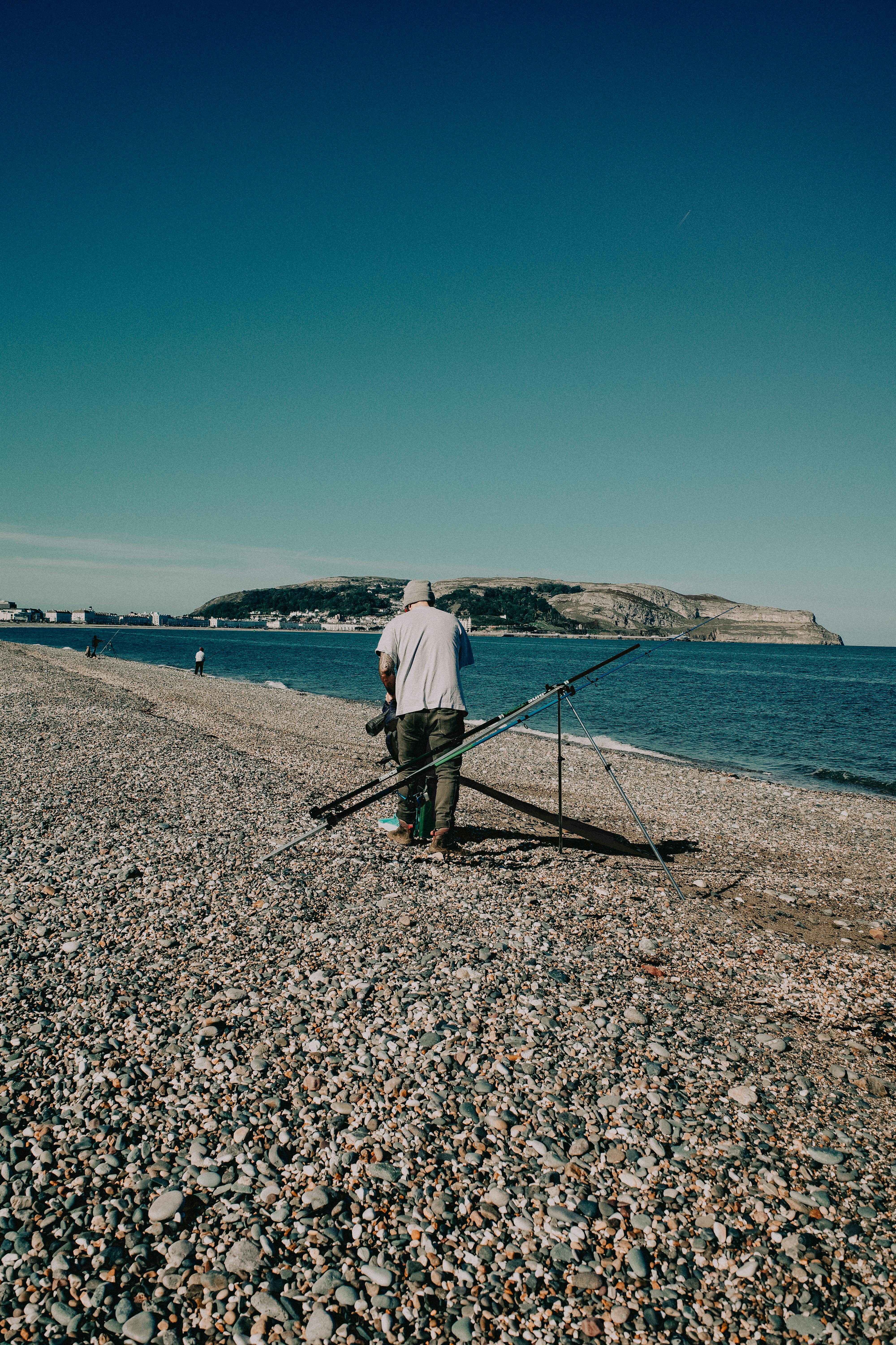 A man fishing on a pebble beach with a scenic view of Llandudno's coastline and blue sky.