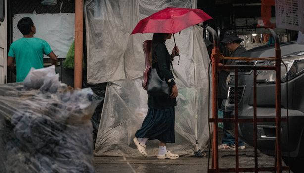 A woman with a red umbrella walks in a rainy Baguio market, carrying a guitar.