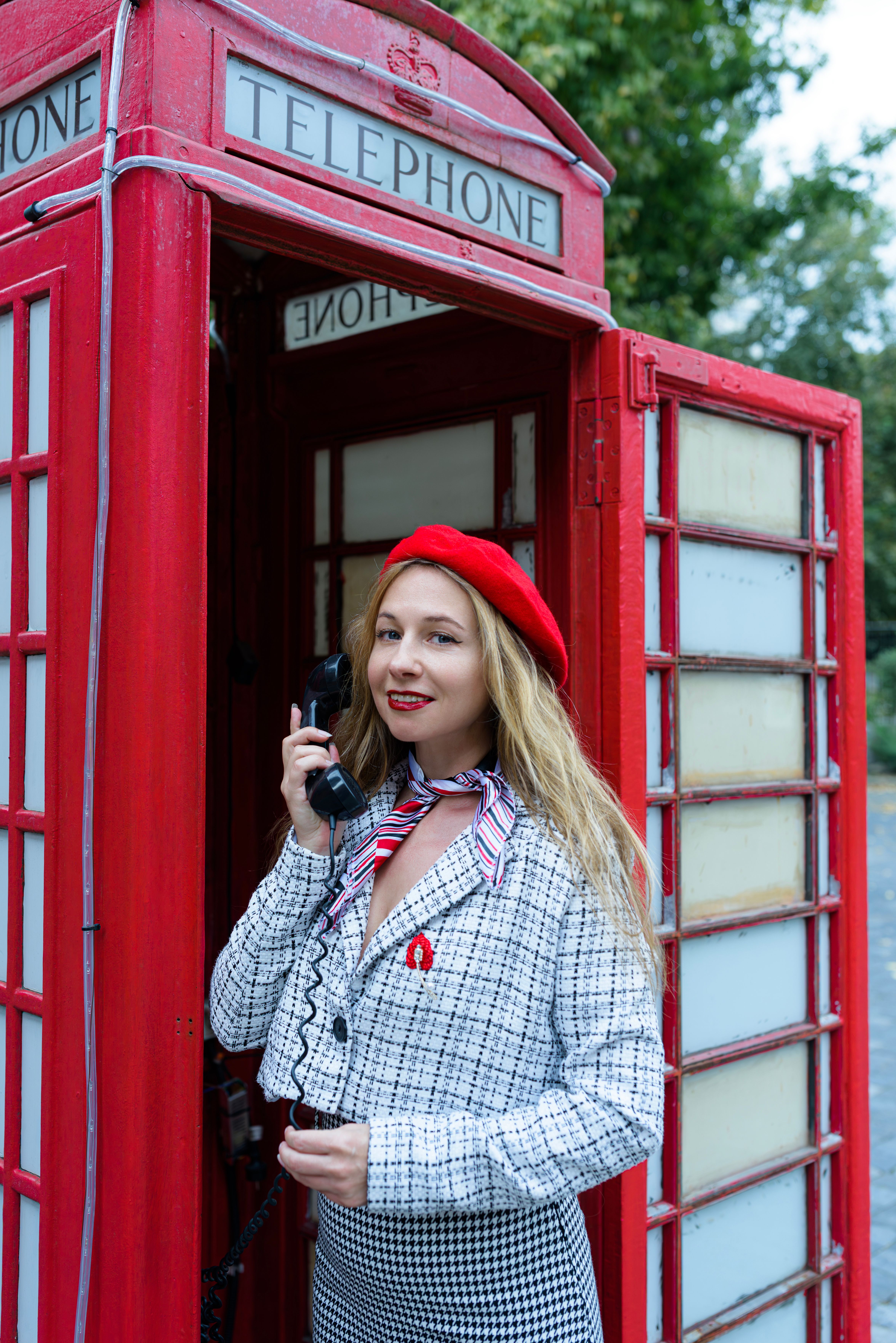Stylish woman in red hat using a vintage British phone booth in an outdoor setting.
