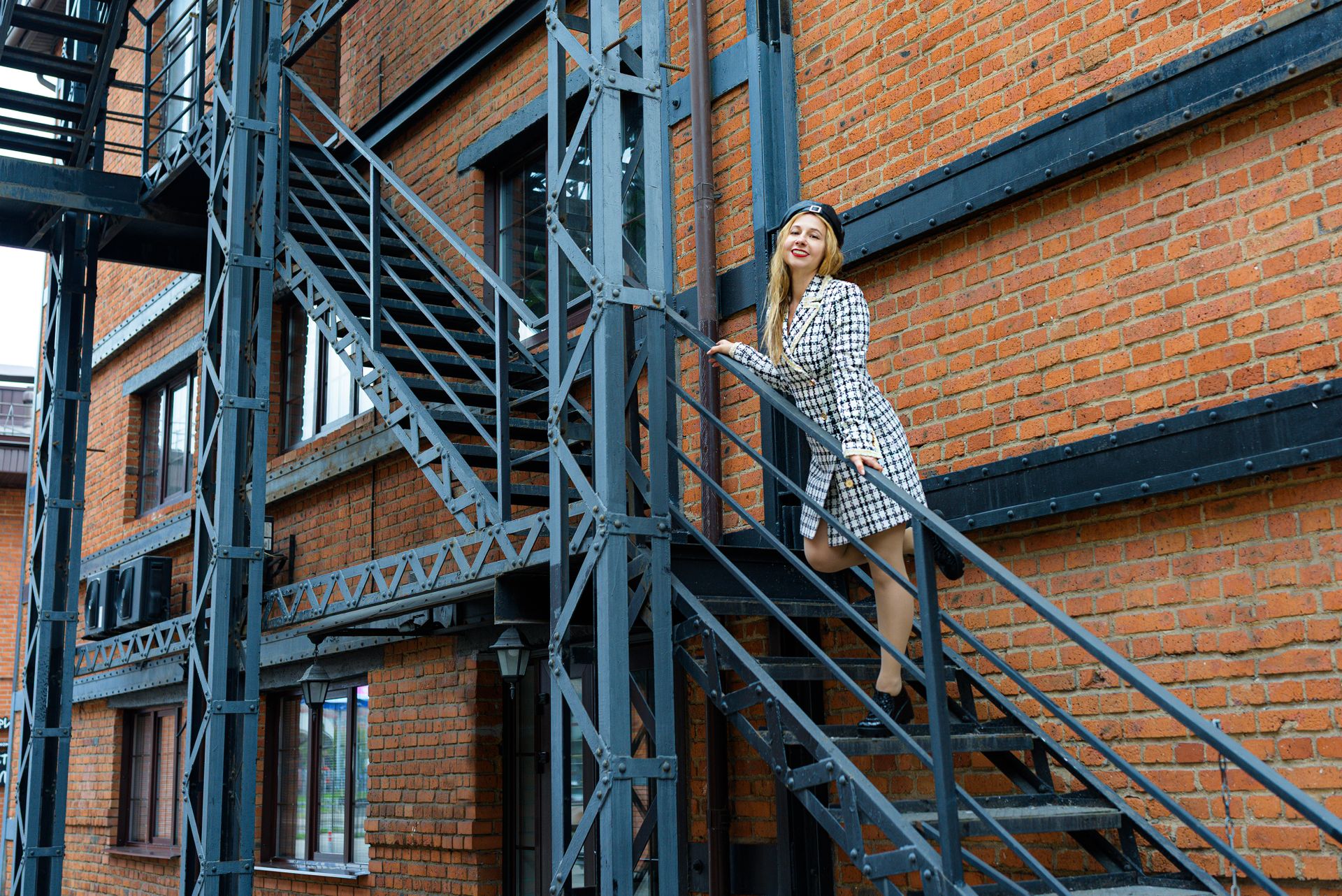 Stylish woman on a metal staircase outside brick building, showcasing urban fashion.