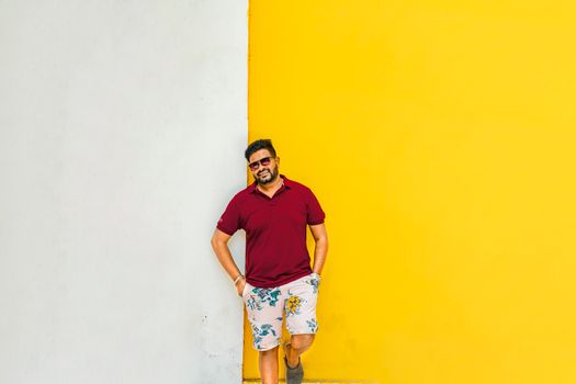 Fashionable adult man in summer attire leans on a vibrant yellow wall in Puducherry, India.
