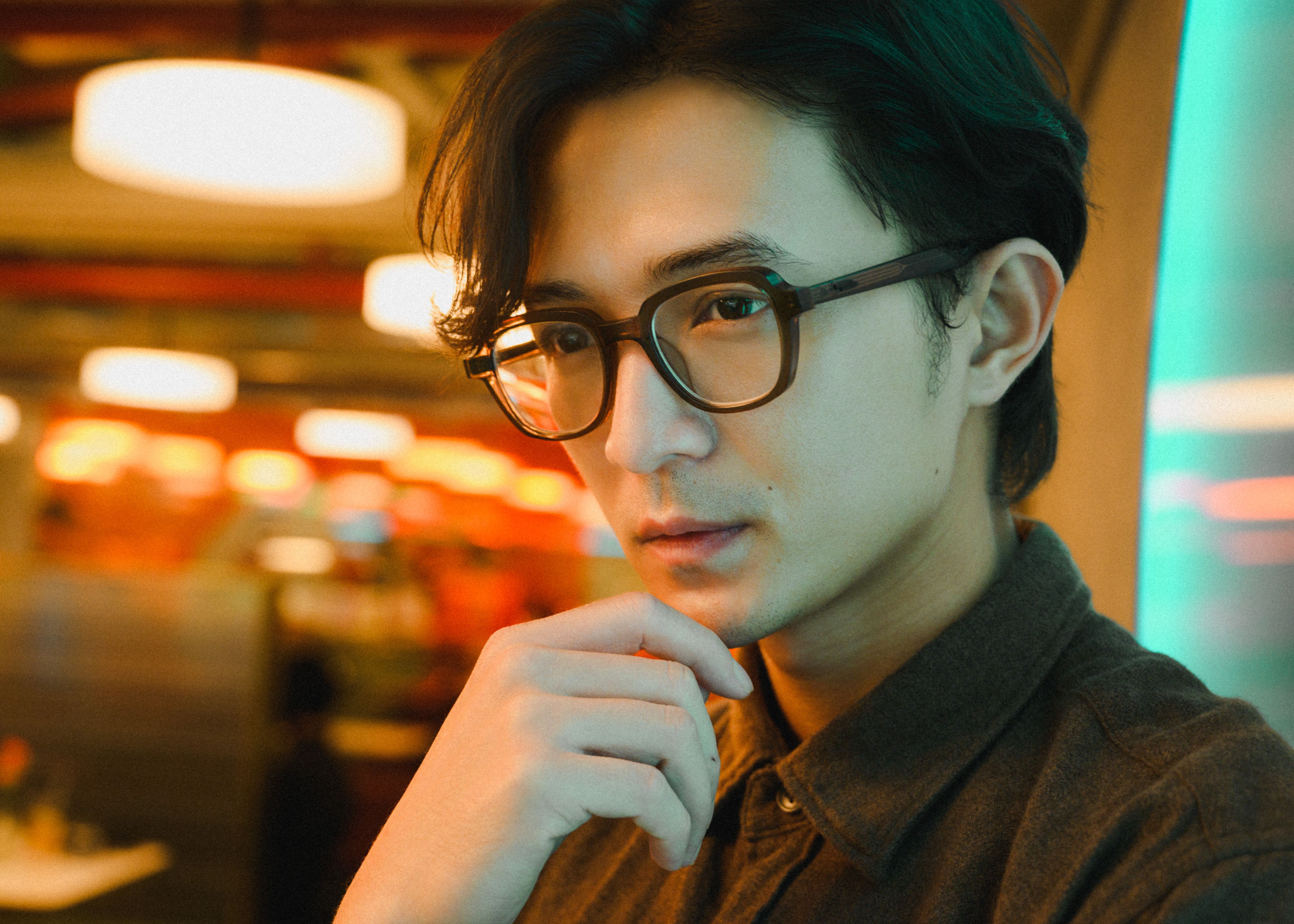 Portrait of a young man with glasses in a modern indoor setting with a thoughtful expression.