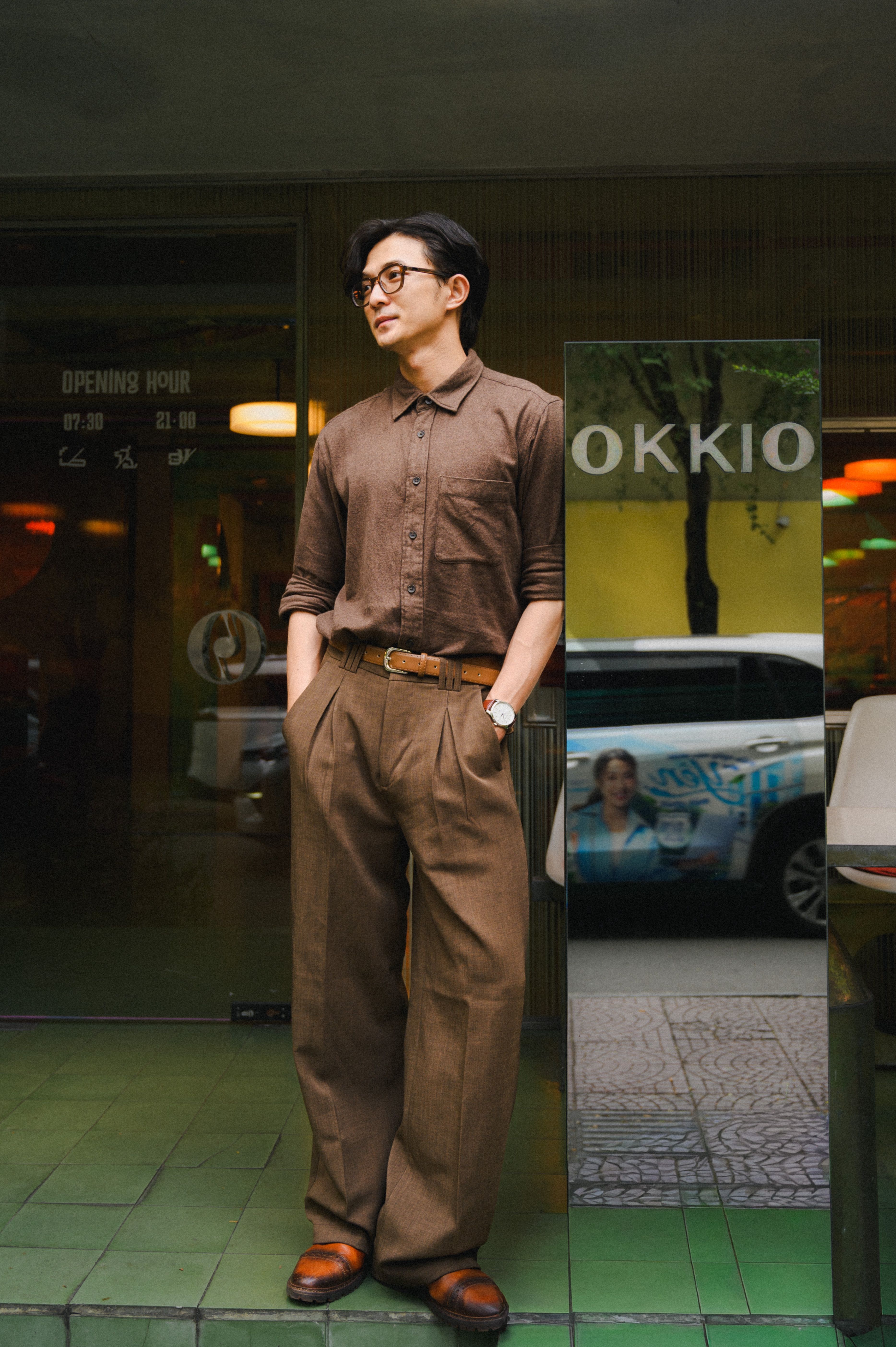 Young man in brown attire stands stylishly outside a cafe with glass reflection.