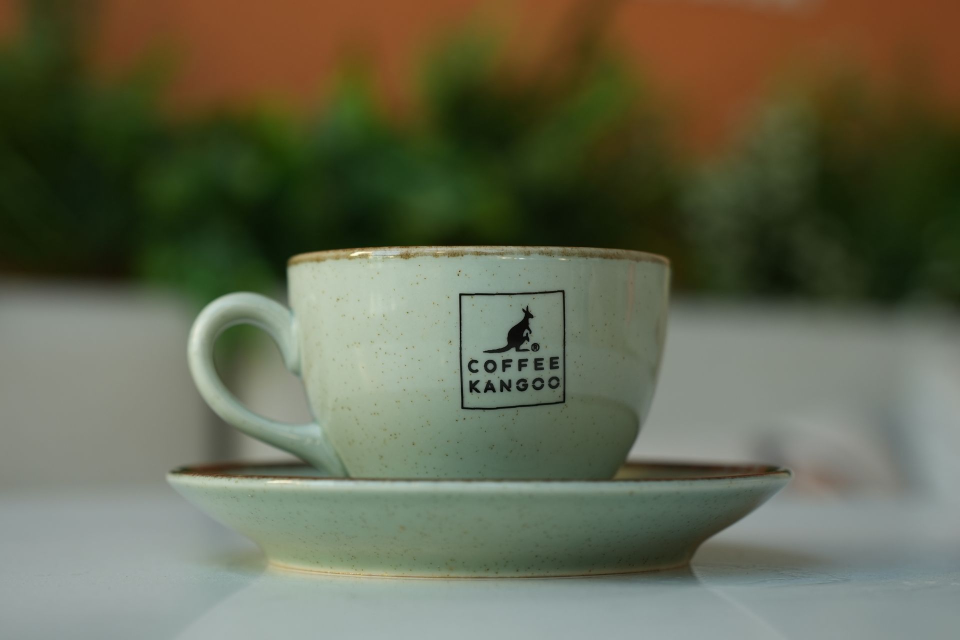 Elegant ceramic coffee cup on saucer with blurred background, featuring stylish 'Coffee Kangoo' logo.