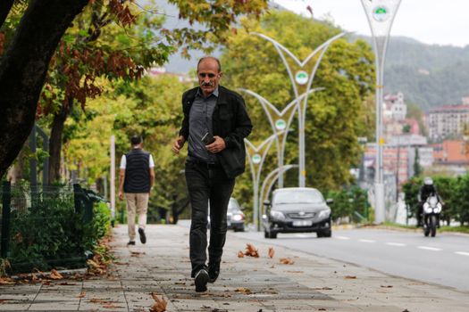 A man runs along a city sidewalk with cars and motorcycles passing by on a fall day.
