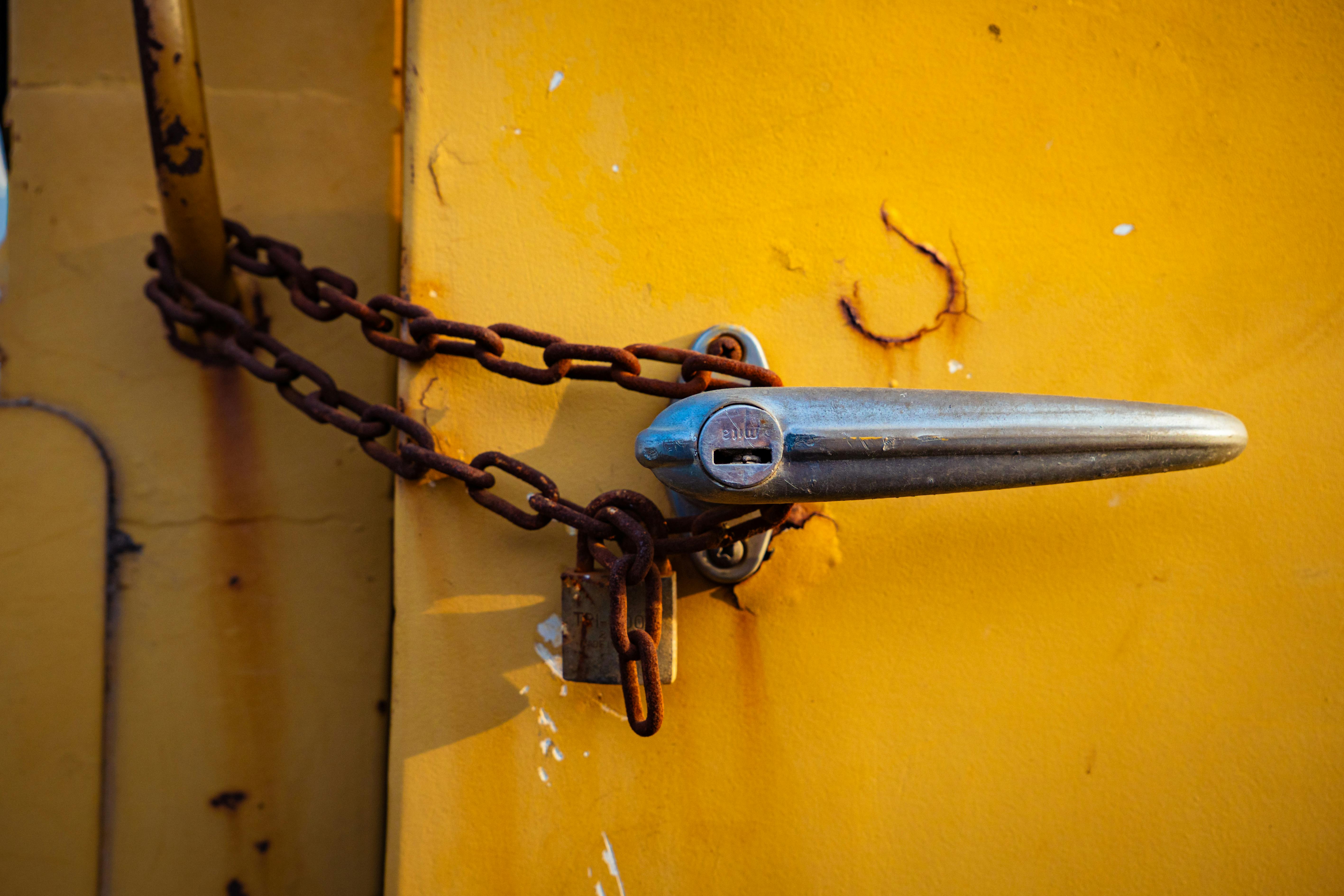 Close-up of a rusty chain securing a chrome handle on an old yellow metal door.