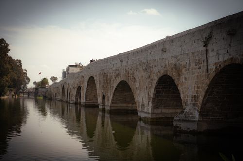 Free Ancient stone bridge arches gracefully over a calm river, surrounded by lush trees. Stock Photo