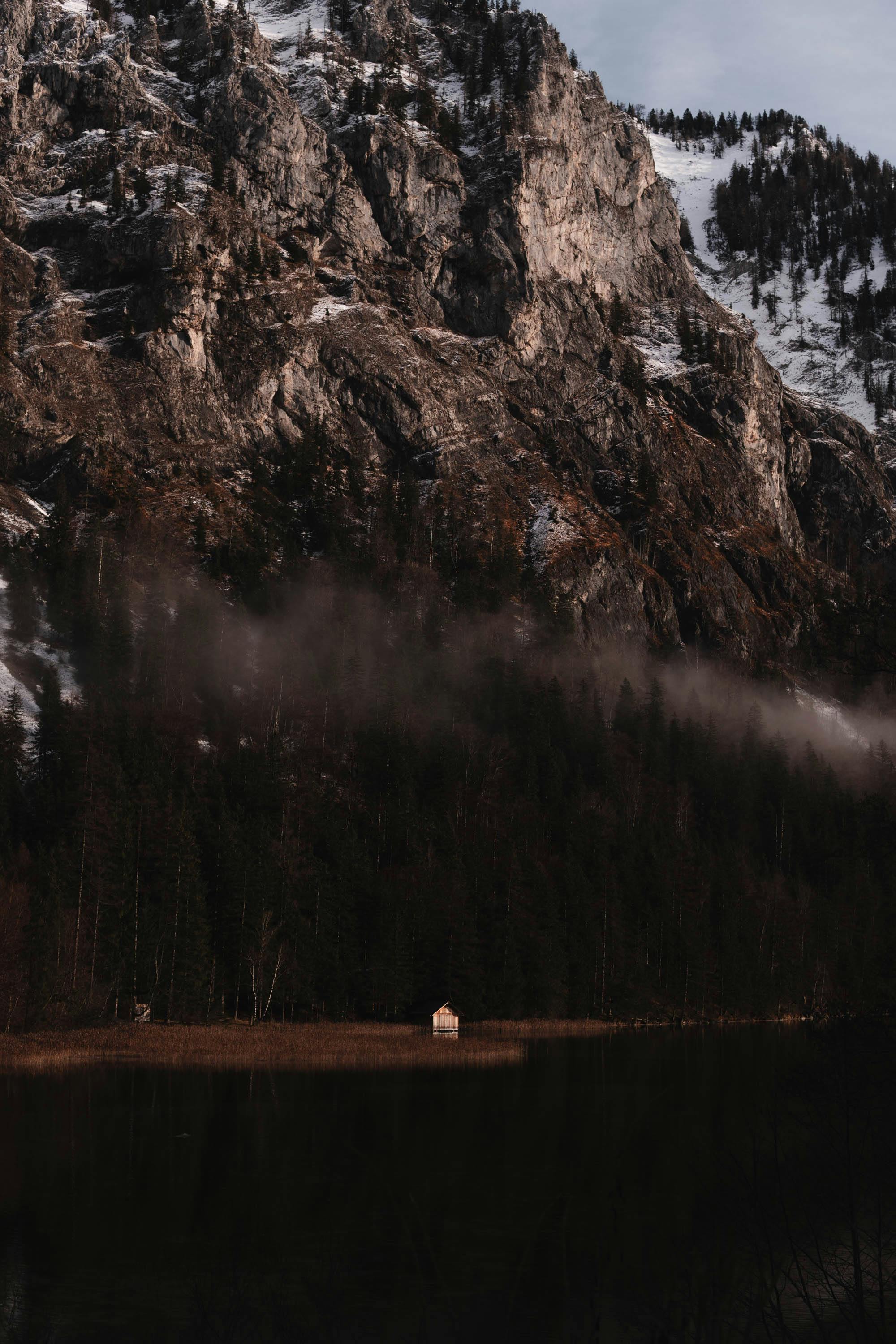 A serene mountain landscape featuring a solitary cabin by the lake with mist and snow-capped peaks.