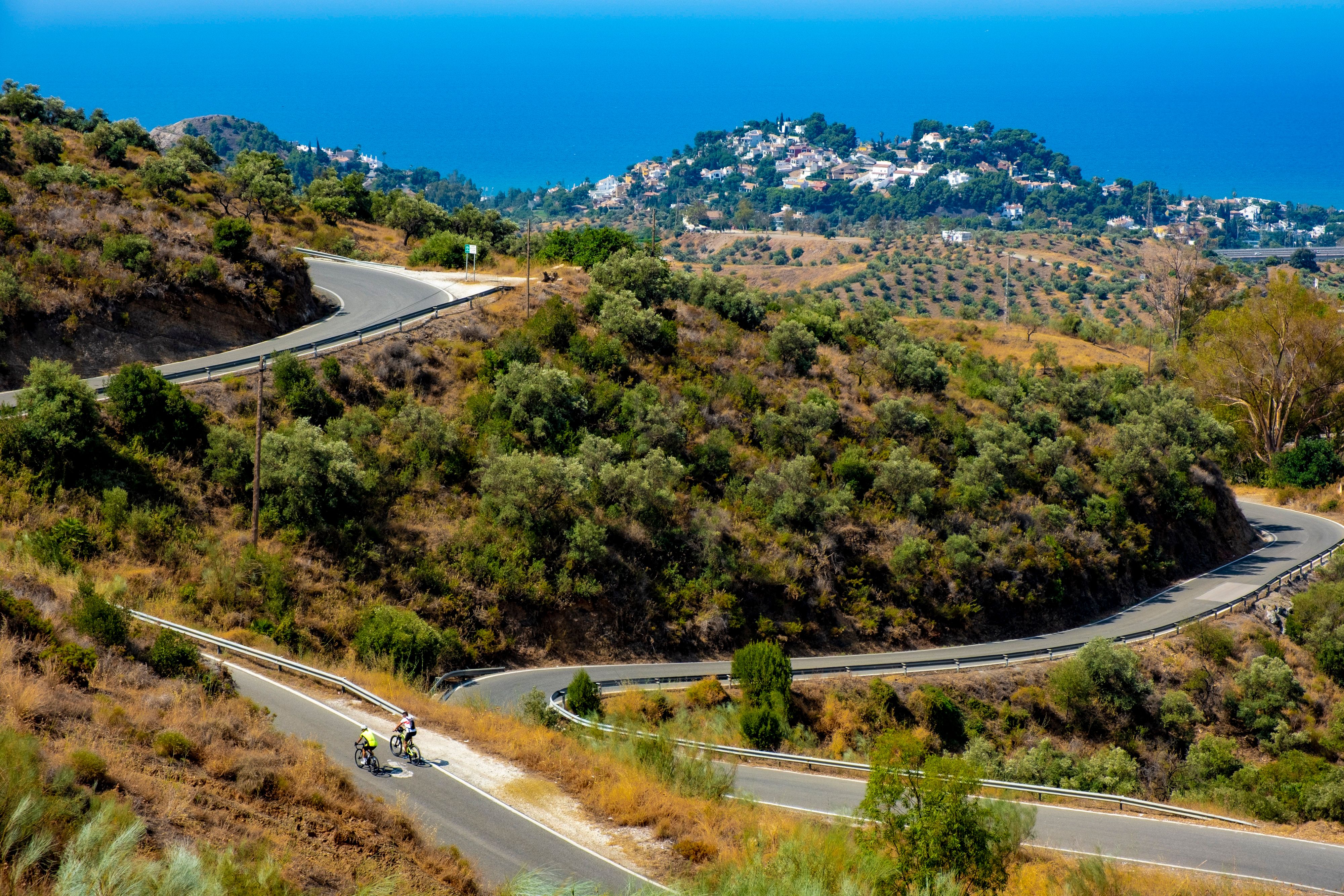 Cyclists on a winding road in Málaga, Spain, surrounded by lush hills and a coastal view.