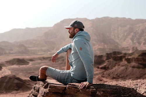 Man in Gray Hoodie and Black Cap Sitting on Brown Rock