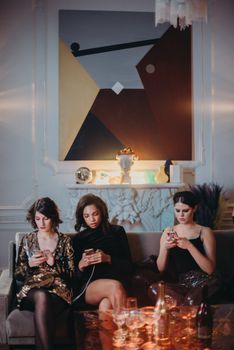Three women in elegant dresses using smartphones at a chic indoor gathering.