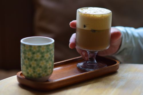 Free Hand holding glass of coffee on a wooden tray with a floral ceramic mug beside it. Stock Photo