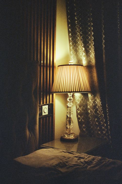 Free Elegant lamp on bedside table illuminating cozy bedroom corner with patterned curtains. Stock Photo