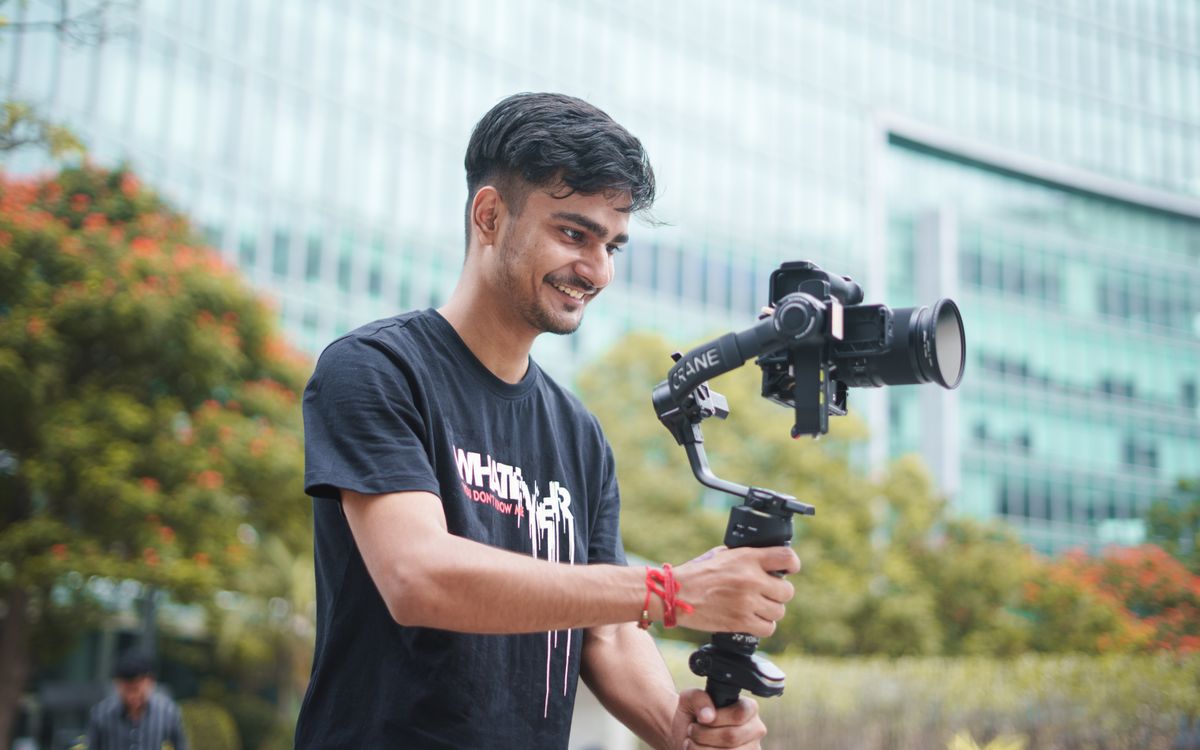 Smiling young videographer using a gimbal outdoors. Urban setting and greenery in the background.